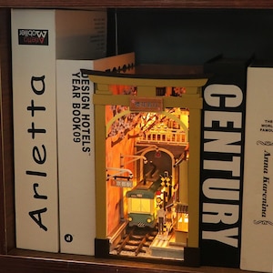 May include: A miniature diorama of a Japanese train station with a cherry blossom tree, a train, and a sign that reads "樱花假日" (Sakura Holiday). The diorama is made of wood and is lit up with LED lights. The diorama is displayed on a bookshelf with other books.