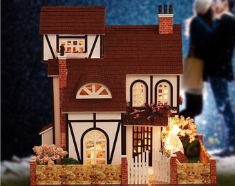May include: A miniature dollhouse with a brown shingled roof, white walls, and black trim. The house has a small front porch with a white picket fence and a small garden with pink flowers. The house is lit up with warm white lights.