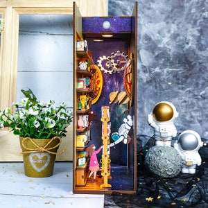 May include: A wooden 3D model of a bookshelf with a galaxy background. The bookshelf is filled with books, a clock, and other decorative items. A small figure of a girl is climbing a ladder to reach a book. The model is lit up with a small light bulb.