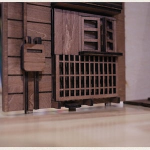 以下が含まれることがあります： 窓とバルコニーのある建物の木製模型。建物は茶色の木でできており、白い背景があります。