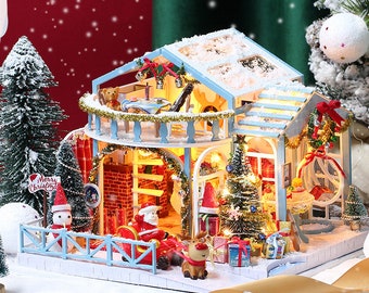 May include: A miniature wooden dollhouse decorated for Christmas. The house has a white roof with a blue base and a balcony with a railing. The house is lit up with lights and has a Christmas tree inside. There is a sign that says "Merry Christmas" in front of the house.