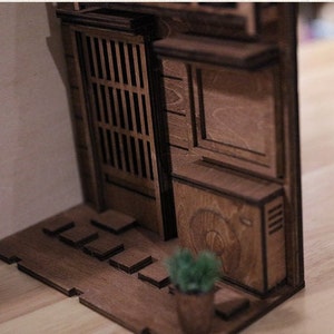 以下が含まれることがあります： 小さなドア、窓、鉢植えの植物が付いた、日本の建物の木製模型。模型は茶色の木でできており、細部までこだわったデザインです。