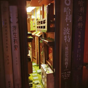 以下が含まれることがあります： 本棚の中に作られた、ミニチュアの街の模型。小さな路地にはレンガの道が敷かれ、バルコニーのある建物や、建物の側面に生えている緑の植物などがある。模型は後ろから光が当たっており、温かい光を放っている。