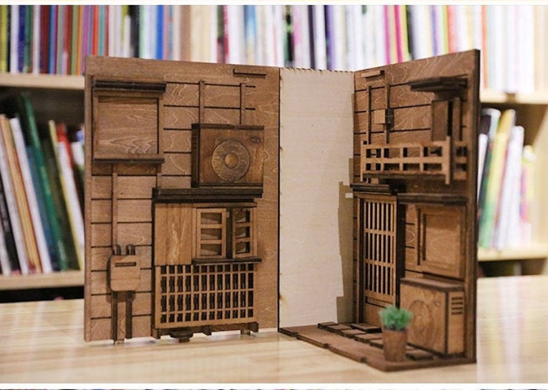 以下が含まれることがあります： レーザーカットされた木材で作られた、複数の建物と小さな鉢植えの植物がある日本の街並みの木製模型。組み立てて楽しむことができます。