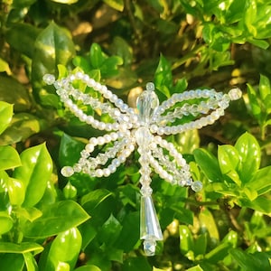 May include: A handcrafted, beaded angel ornament with clear and iridescent beads. The angel features pearl-like beads for the wings and body, and a clear, faceted crystal for the head and skirt. The ornament is set against a backdrop of green foliage.