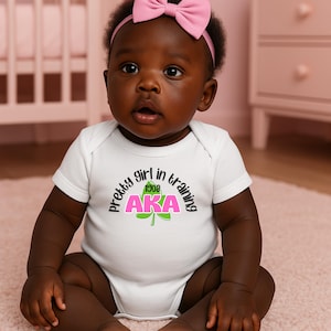 May include: A baby wearing a white onesie with the text "pretty girl in training" above the letters "AKA" in pink and green. The baby has a pink bow headband.