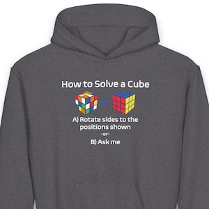 Op de afbeelding: Een donkergrijs hoodie met de tekst "How to Solve a Cube" en een afbeelding van een Rubik's Cube. De afbeelding toont twee Rubik's Cubes, een met gemengde kleuren en de andere met opgeloste kleuren. De tekst onder de afbeelding luidt "A) Rotate sides to the positions shown -or- B) Ask me."