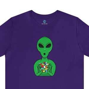 May include: Purple t-shirt with a green alien holding a colorful Rubik's Cube.