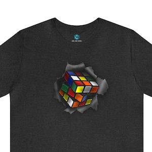 May include: A dark grey t-shirt with a colorful Rubik's Cube breaking through the fabric. The cube is in a 3D style and appears to be popping out of the shirt.