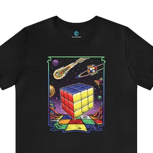 Puede incluir: Una camiseta negra con un gráfico colorido de un cubo de Rubik en el espacio. El cubo flota en una galaxia con un cometa y planetas. El gráfico está rodeado por un marco negro.