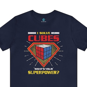 Op de afbeelding: Donkerblauw T-shirt met een rode, witte en blauwe Rubik's Cube in een geel superheldenlogo. De tekst "I Solve Cubes" staat boven het logo en "What's Your Superpower?" staat onder het logo.