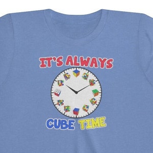 Puede incluir: Una camiseta azul claro para jóvenes con un gráfico de una esfera de reloj con cubos de Rubik como números. El texto "IT'S ALWAYS CUBE TIME" está impreso en la camiseta.