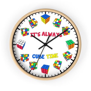 Op de afbeelding: Een witte ronde klok met een houten frame. De wijzerplaat van de klok is zwart-wit met zwarte wijzers. De wijzerplaat van de klok is versierd met kleurrijke Rubik's Cubes rond de wijzerplaat. De tekst "It's Always Cube Time" is gedrukt op de wijzerplaat van de klok.