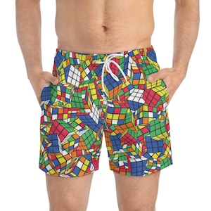 Puede incluir: Pantalones cortos de baño con un colorido diseño de Cubo de Rubik. Los pantalones cortos tienen cintura con cordón y bolsillos. El diseño incluye cubos rojos, azules, verdes, amarillos y naranjas sobre un fondo blanco.