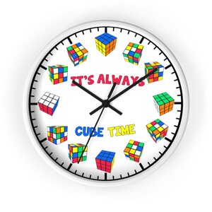 Op de afbeelding: Witte ronde klok met zwarte wijzers en een witte achtergrond. De wijzerplaat heeft een zwarte rand en zwarte markeringen voor de uren en minuten. De wijzerplaat heeft ook een kleurrijk Rubik's Cube-ontwerp met de tekst "It's Always Cube Time".