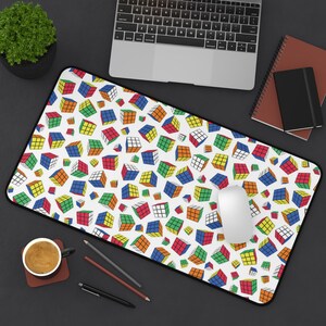May include: A rectangular mouse mat with a black border displays a repeating pattern of colourful Rubik's Cubes on a white background. A white computer mouse sits on the mat, with a laptop and other office supplies in the background.
