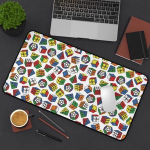 May include: A rectangular white mousepad with a black border, covered in a colorful pattern of Rubik's Cubes and other geometric puzzles. A computer mouse sits on the pad, with a laptop, notebook, and coffee cup in the background.