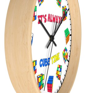 Op de afbeelding: Een ronde houten klok met een witte wijzerplaat en zwarte wijzers. De wijzerplaat van de klok heeft een kleurrijk Rubik's Cube-ontwerp met de tekst "It's Always Cube Time".