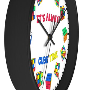 Op de afbeelding: Een zwart-witte klok met een witte wijzerplaat en zwarte wijzers. De wijzerplaat van de klok heeft een kleurrijk ontwerp van Rubik's Cubes en de tekst "It's Always Cube Time".
