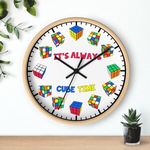 Op de afbeelding: Een witte klok met zwarte wijzers en een houten frame. De wijzerplaat van de klok heeft een kleurrijk Rubik's Cube-ontwerp met de tekst "It's Always Cube Time".
