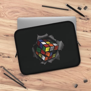 May include: Black laptop sleeve with a colourful Rubik's Cube design bursting through a torn paper effect. The sleeve has a zip closure and is on a wooden surface with pencils and a pen.