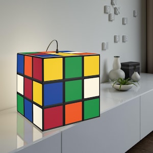 以下が含まれることがあります： 黒いフレームの立方体型ランプ。ルービックキューブのデザインで、赤、オレンジ、黄色、緑、青、白の正方形があります。上部から黒いコードが出ています。ランプは白い表面に置かれています。