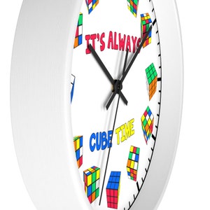 Op de afbeelding: Witte ronde klok met zwarte wijzers en een witte achtergrond. De wijzerplaat van de klok heeft een kleurrijk patroon van Rubik's Cubes en de tekst "It's Always Cube Time".
