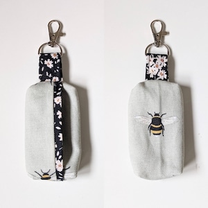 以下が含まれることがあります： 薄い緑色の布製のポーチと白黒の花柄のストラップが付いたキーホルダーが2つ。1つのキーホルダーには、ポーチに蜂が刺繍されています。