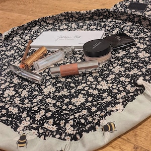 May include: A black floral makeup bag with a selection of cosmetics. The bag contains items such as eyeshadow palettes, lipsticks, and a powder compact. The bag's trim has a light green border with bee illustrations.