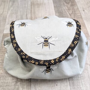 May include: A light blue cosmetic bag with a flap closure, adorned with embroidered bees. The flap is trimmed with a black and gold floral pattern. The bag is made of a soft, textured fabric.