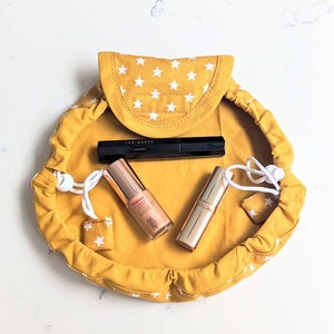 May include: A yellow drawstring makeup bag with white stars printed on it. The bag is open and contains three makeup items: a black mascara, a gold lipstick, and a gold foundation.