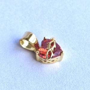 Può includere: Ciondolo in oro con una pietra preziosa rosso-arancio a forma di cuore. Il ciondolo ha un'ansa dorata decorativa e un'incastonatura dorata dettagliata. La pietra preziosa è sfaccettata, riflettendo la luce. Il gioiello è posto su uno sfondo chiaro.