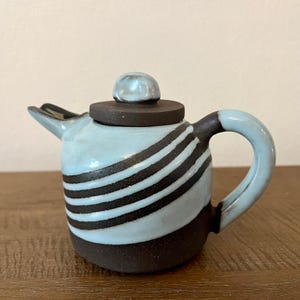 May include: A small, ceramic teapot with a light blue glaze and dark brown stripes. The teapot has a matching lid and handle, and is set on a wooden surface. The teapot is a unique piece of pottery.