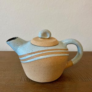 May include: A ceramic teapot with a light blue speckled glaze and tan base. The teapot features horizontal stripes and a matching lid with a small knob. The handle and spout are also light blue.