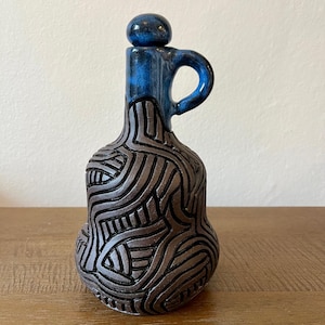 May include: A ceramic bottle with a dark blue handle and stopper. The bottle's body is brown with a textured, carved design. The handle and top portion are a glossy dark blue. The bottle sits on a wooden surface.