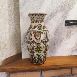 May include: A brown and white ceramic vase with a floral pattern in shades of green, yellow, and brown. The vase has a wide mouth and a narrow base.