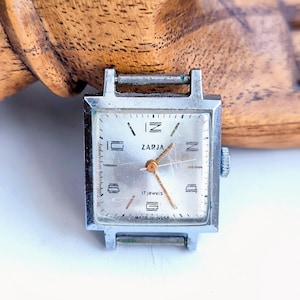 Könnte beinhalten: Vintage ZARJA Armbanduhr in Silber mit rechteckigem Zifferblatt. Das Zifferblatt ist silberfarben mit schwarzen Ziffern und Zeigern. Die Uhr zeigt "17 jewels" und "Made in USSR". Die Uhr ist vor einem Holzuntergrund platziert.