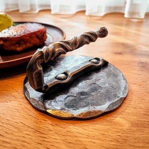 May include: A handcrafted, dark metal burger press with a round base and a twisted handle. The base has a hammered texture and a decorative edge. The handle is curved and has a detailed design.