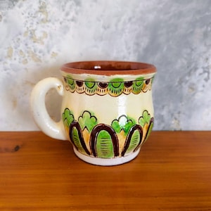 May include: A small, white ceramic mug with a brown rim and green and brown painted designs. The mug has a rounded body and a handle.