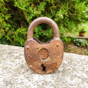 May include: A rusty padlock with a heart-shaped design and the text "JOHN KENT & SONS" on the front. The padlock is sitting on a grey concrete surface.