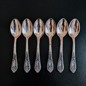 May include: Six silver spoons with ornate floral designs on the handles are arranged on a black surface. The spoons have a reflective, polished finish, and the handles feature detailed engravings.