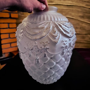 May include: A frosted glass decorative lamp shade with a quilted pattern on the bottom and ornate draped and tassel details. The top has a ribbed design. The shade is held by a hand.