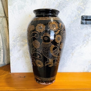 May include: A black ceramic vase with a glossy finish, adorned with gold-colored floral and bird motifs. The vase has a wide base, a cylindrical body, and a slightly flared rim. The design features detailed patterns.