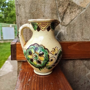 May include: A white ceramic pitcher with green, yellow, and brown floral designs. The pitcher has a handle and a spout.