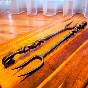 May include: Two hand-forged metal carving forks with a dark, textured finish. Each fork features a long, slender handle with a decorative twisted metal design and a curved, scrolled end. The two-pronged heads are pointed and functional.