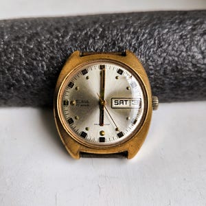 May include: A vintage gold-coloured wristwatch with a silver face. The watch face displays the day of the week as "SAT" and the brand name "SLAVA". The watch has a black leather strap and a gold-coloured bezel.