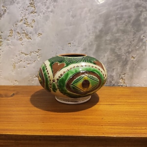 May include: A small, round, green and brown ceramic vase with a decorative pattern. The vase has a white base and is sitting on a wooden surface.
