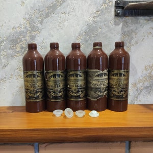Puede incluir: Cinco botellas de vidrio marrón con etiquetas doradas y texto negro que dice "RIGAS MELNAIS BALZAMS" y "ЧЕРНЫЙ БАЛЬЗАМ". Las botellas están dispuestas en una fila sobre una superficie de madera. Hay cinco tapones de botella de plástico blanco delante de las botellas.