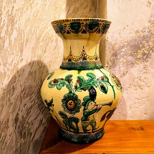 May include: A decorative ceramic vase with a wide base and flared top. The vase is painted with a yellow background and features green and blue floral and figure designs. The rim and base have a dark green trim.