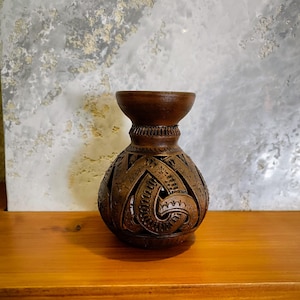 May include: A brown, carved ceramic vase with a wide base and a narrow neck. The vase features intricate cut-out patterns and a textured surface. The vase is displayed on a wooden surface against a neutral background.